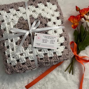 May include: A hand-crocheted granny square throw blanket in shades of dusty rose and white. The blanket is tied with a white ribbon and a tag that reads "I'm hand crocheted. Here is how to take extra care of me."
