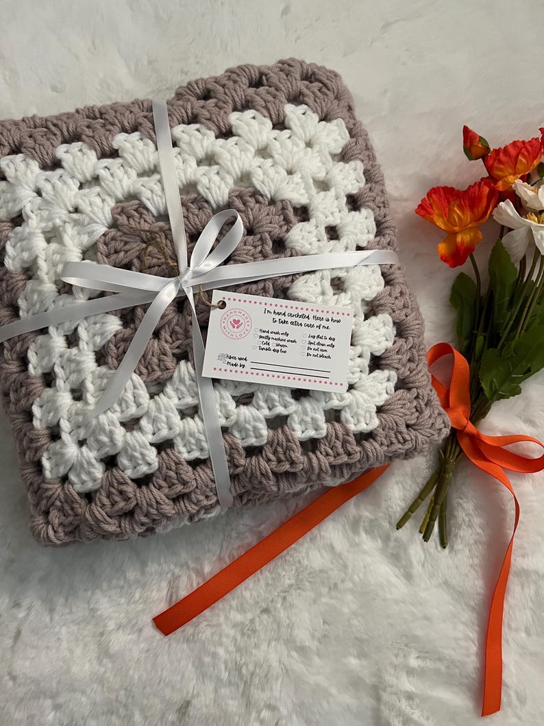 May include: A hand-crocheted granny square throw blanket in shades of dusty rose and white. The blanket is tied with a white ribbon and a tag that reads "I'm hand crocheted. Here is how to take extra care of me."