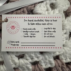 May include: A white tag with pink polka dots attached to a crocheted blanket with pink and white yarn. The tag says "I'm hand crocheted. Here is how to take extra care of me." and lists instructions for washing and drying the blanket. The tag also has a space to write the fiber used and who made the blanket.