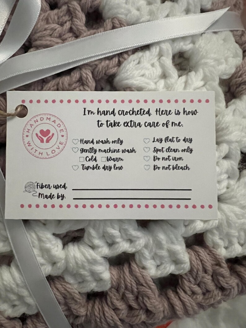 May include: A white tag with pink polka dots attached to a crocheted blanket with pink and white yarn. The tag says "I'm hand crocheted. Here is how to take extra care of me." and lists instructions for washing and drying the blanket. The tag also has a space to write the fiber used and who made the blanket.