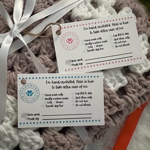 May include: A pink and white crocheted blanket with a care tag attached. The tag says "I'm hand crocheted. Here is how to take extra care of me." and lists instructions for washing and drying the blanket. The tag also has a space to write in the fiber used and who made the blanket.