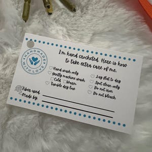May include: White card with blue and black text and a heart logo that says "Handmade with Love". The card lists care instructions for a crocheted item, including hand washing, gentle machine washing, tumble drying low, laying flat to dry, spot cleaning, and not ironing or bleaching.