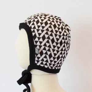 May include: Black and white geometric patterned baby bonnet with a black tie. The bonnet has a black trim around the edge.