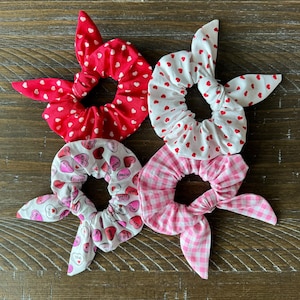 May include: Four fabric scrunchies with bow accents. One is red with white hearts, another is white with red hearts, a third is white with candy heart graphics, and the last is pink gingham.