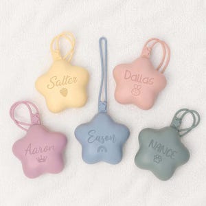 Può includere: Cinque portachiavi in silicone a forma di stella in colori pastello: giallo, rosa, blu, malva e verde salvia. Ogni portachiavi presenta un occhiello, un nome personalizzato e una piccola icona: fragola, ape, corona, arcobaleno e zampa.