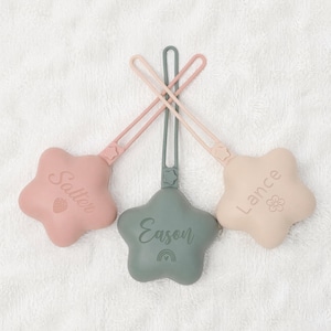 May include: Three star-shaped silicone dummy clips with long straps. The left clip is dusty pink with the name Salter and a strawberry icon. The centre clip is sage green with the name Eason and a rainbow icon. The right clip is beige with the name Lance and a flower icon.