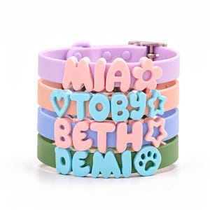 Op de afbeelding: Een stapel van vier siliconen armbanden in lavendel, perzik, maagdenpalm en olijfgroen. Elke armband heeft verhoogde, kleurrijke letters in bubbelstijl die de namen MIA, TOBY, BETH en DEMI spellen, vergezeld van kleine bloem-, hart- en pooticonen.
