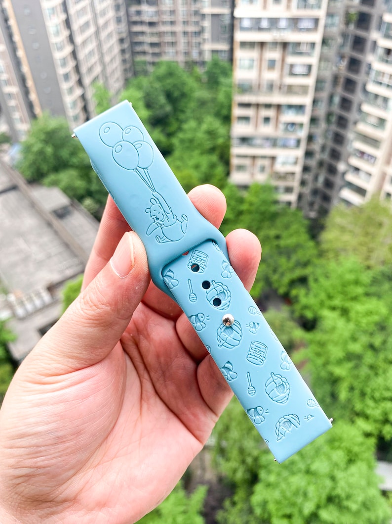May include: Light blue silicone watch band with a Winnie the Pooh design. The band features a cartoon image of Winnie the Pooh holding balloons and a pattern of honey pots, bees, and honeycombs.