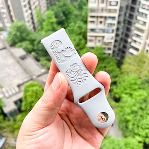 May include: A light gray watch band with engraved images of the Cheshire Cat from Alice in Wonderland. The band has a silver metal clasp and is held in a hand against a background of green trees and buildings.