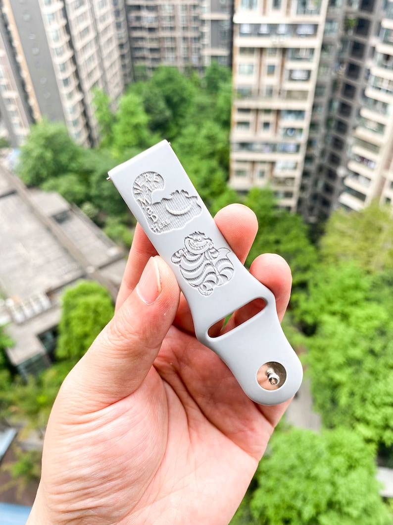 May include: A light gray watch band with engraved images of the Cheshire Cat from Alice in Wonderland. The band has a silver metal clasp and is held in a hand against a background of green trees and buildings.