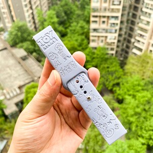 以下が含まれることがあります： トイストーリーのデザインが施されたライトグレーのシリコン製腕時計バンド。バンドにはウッディ、バズ・ライトイヤー、その他のトイストーリーのキャラクターが描かれています。バンドには「トイストーリー」の文字が印刷されています。