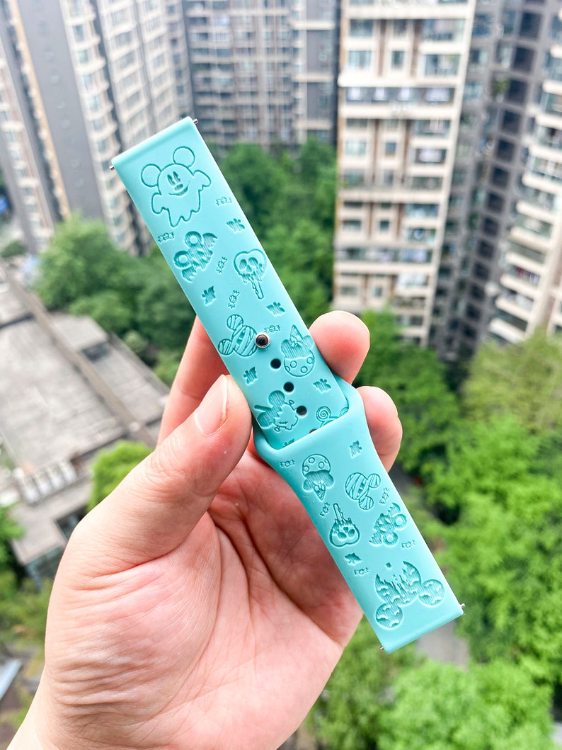 May include: A light blue silicone watch band with a variety of embossed cartoon characters, including Mickey Mouse, ghosts, and pumpkins. The band is designed for a smartwatch.