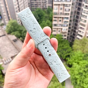 May include: A light blue silicone watch strap with a repeating pattern of embossed cartoon characters. The strap is designed for a smartwatch.