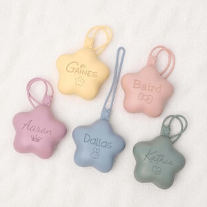 Peut inclure: Cinq porte-clés en silicone en forme d'étoile aux couleurs pastel, chacun portant un nom et une petite icône. Le jaune indique Gaines avec une étoile, le rose Baird avec un nœud, le violet Aaron avec une couronne, le bleu Dallas avec une abeille et le vert Kathie avec des cœurs.