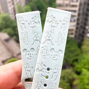 Peut inclure: Un bracelet de montre en silicone bleu clair avec un motif répété de visages de personnages Disney blancs, notamment Mickey Mouse, Minnie Mouse, Donald Duck et Dingo.