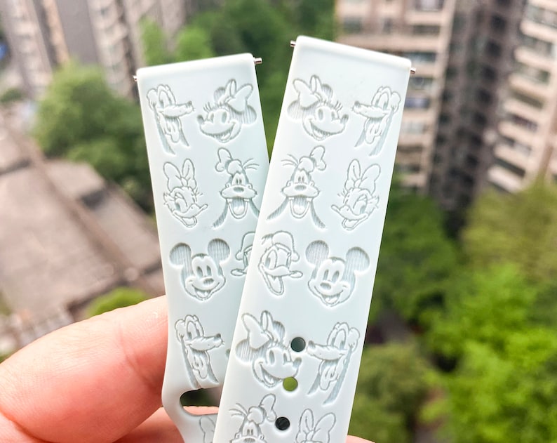 May include: A light blue silicone watch strap with a repeating pattern of white Disney character faces, including Mickey Mouse, Minnie Mouse, Donald Duck, and Goofy.