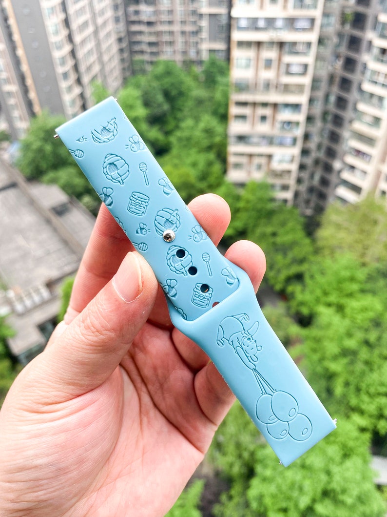 May include: A light blue silicone watch band with a Winnie the Pooh design. The band features a repeating pattern of honey pots, bees, and Winnie the Pooh holding balloons.