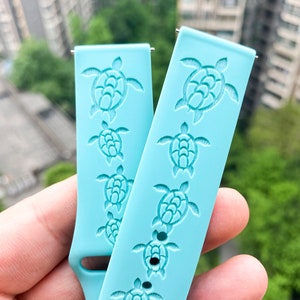 Puede incluir: Dos correas de reloj de silicona azul claro con un patr&oacute;n repetido de tortugas marinas. Las tortugas est&aacute;n en relieve en las correas.