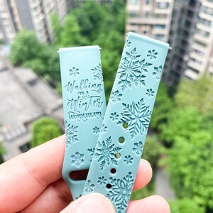 Op de afbeelding: Twee lichtblauwe horlogebandjes met een sneeuwvlokkenpatroon. Een bandje heeft de tekst "Walking in a Winter Wonderland" erop gedrukt.
