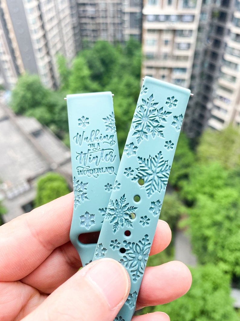 Op de afbeelding: Twee lichtblauwe horlogebandjes met een sneeuwvlokkenpatroon. Een bandje heeft de tekst "Walking in a Winter Wonderland" erop gedrukt.