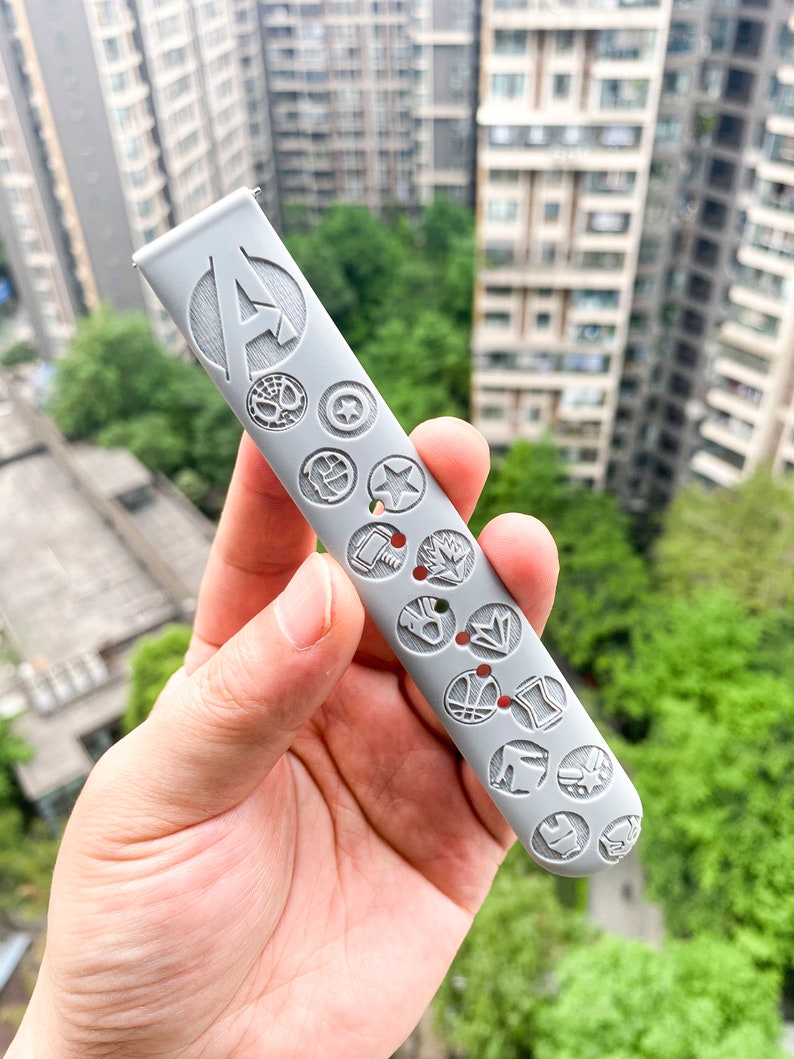 May include: A gray silicone watch band with a raised design of various Marvel characters, including Iron Man, Captain America, Thor, and Spider-Man. The band has the letter "A" for Avengers in the center.