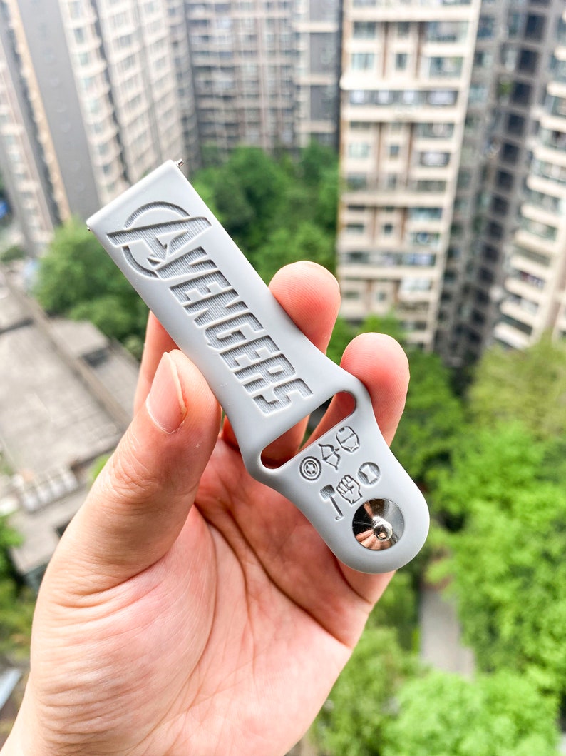 May include: A gray silicone watch band with the word "Avengers" embossed on it. The band has a silver metal buckle with various superhero symbols embossed on it.
