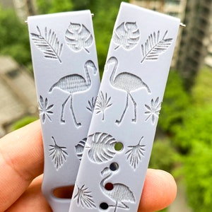 Puede incluir: Dos correas de reloj de silicona gris claro con un tema tropical. Las correas presentan un patr&oacute;n repetitivo de siluetas blancas de flamencos y hojas de palmeras.