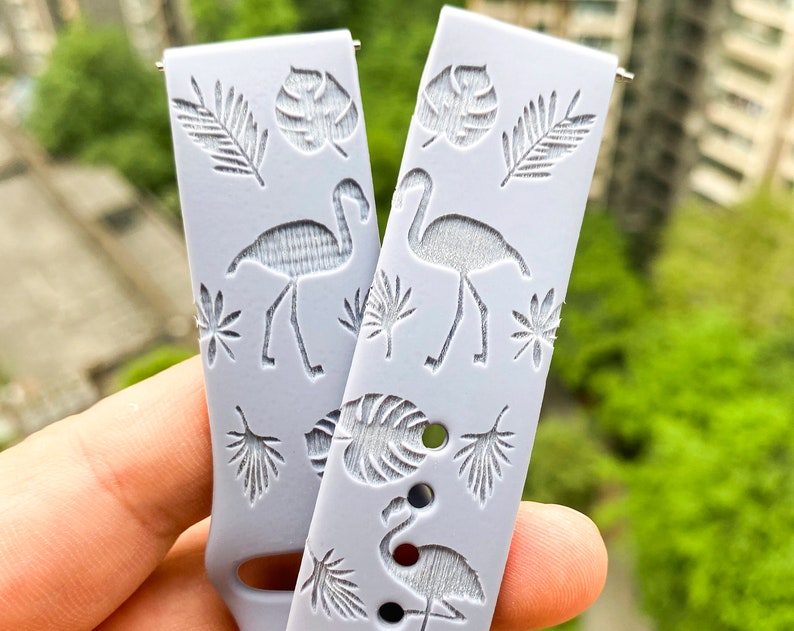 Puede incluir: Dos correas de reloj de silicona gris claro con un tema tropical. Las correas presentan un patr&oacute;n repetitivo de siluetas blancas de flamencos y hojas de palmeras.
