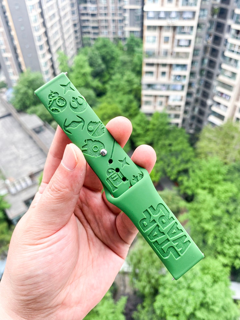 May include: Green silicone watch strap with a Star Wars theme. The strap features various Star Wars characters and symbols, including R2-D2, Darth Vader, and the Millennium Falcon. The strap also has the words "Star Wars" embossed on it.