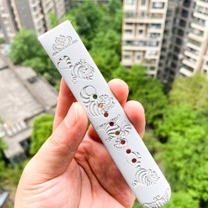 May include: A light gray watch band featuring engraved images of the Cheshire Cat from Alice in Wonderland. The band has a series of the cat's smiling faces and striped bodies. The band has several holes for adjustment and two metal pins for attachment.