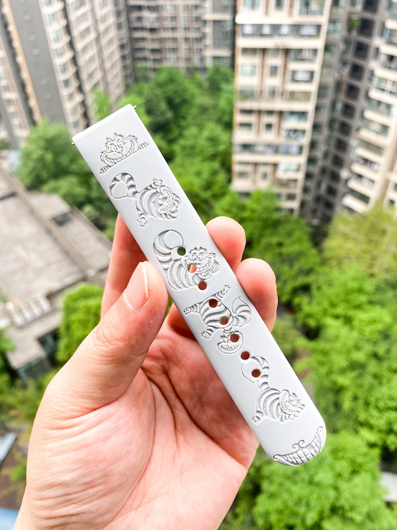 May include: A light gray watch band featuring engraved images of the Cheshire Cat from Alice in Wonderland. The band has a series of the cat's smiling faces and striped bodies. The band has several holes for adjustment and two metal pins for attachment.