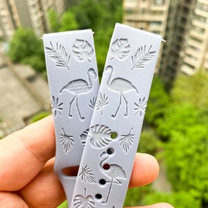 Puede incluir: Una correa de reloj de silicona gris con un estampado tropical con flamencos y hojas de palma.