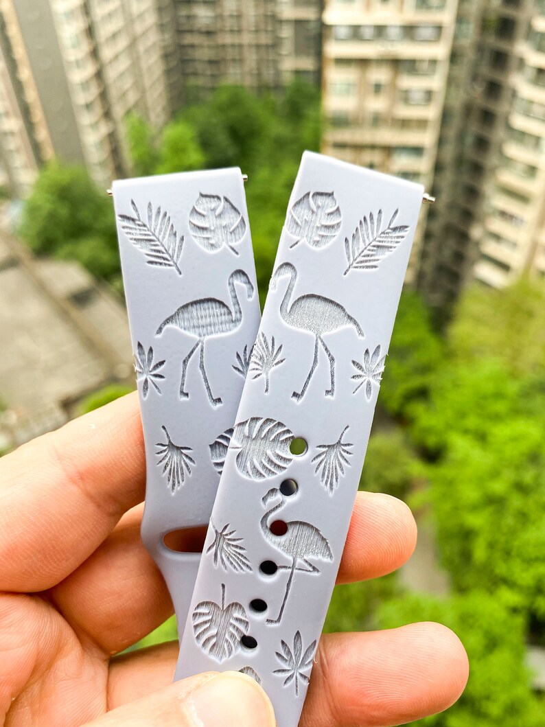 Puede incluir: Una correa de reloj de silicona gris con un estampado tropical con flamencos y hojas de palma.