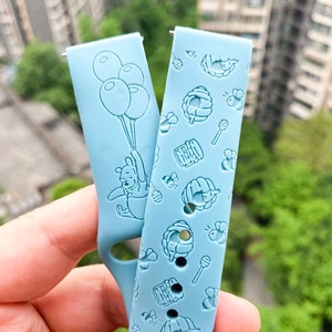 May include: Two light blue silicone watch bands with embossed designs. One band features Winnie the Pooh holding balloons, the other has a repeating pattern of honey pots, bees, and honey sticks.