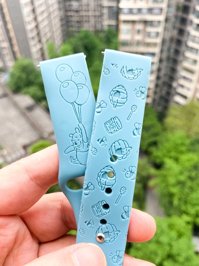 May include: Two light blue silicone watch bands with embossed designs. One band features Winnie the Pooh holding balloons, the other has a repeating pattern of honey pots, bees, and honey sticks.