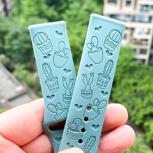 Puede incluir: Dos correas de reloj de silicona azul claro con un dise&ntilde;o de cactus y maceta. Las correas tienen un patr&oacute;n repetitivo de cactus en macetas con corazones.