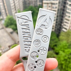 May include: Two gray silicone watch bands with embossed Avengers logos and character symbols. The band on the left has the word "Avengers" embossed on it. The band on the right has the Avengers "A" logo embossed on it.