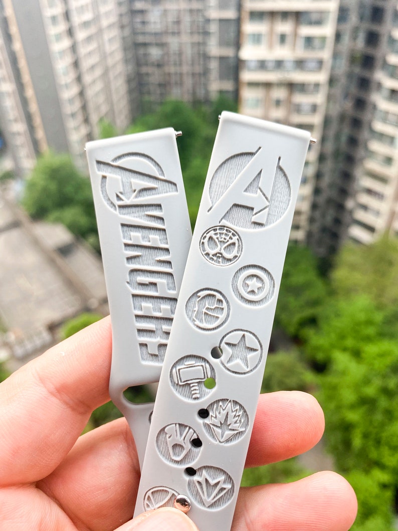 May include: Two gray silicone watch bands with embossed Avengers logos and character symbols. The band on the left has the word "Avengers" embossed on it. The band on the right has the Avengers "A" logo embossed on it.