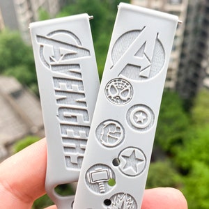 May include: Two white watch bands with embossed designs. One band features the word "Avengers" and the other features the letter "A" in a circle with other Marvel character symbols.
