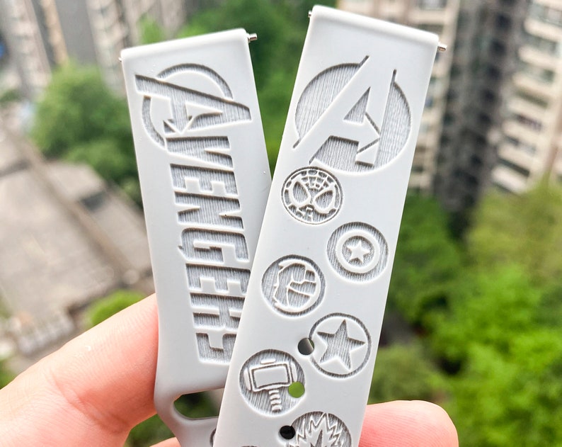 May include: Two white watch bands with embossed designs. One band features the word "Avengers" and the other features the letter "A" in a circle with other Marvel character symbols.