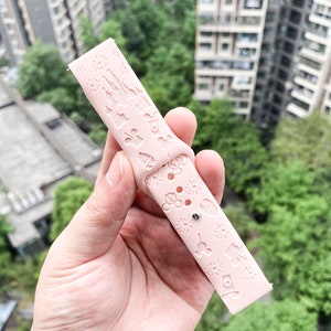 May include: A pink silicone watch band with a white embossed design featuring Disney characters and landmarks. The band has a small silver buckle.