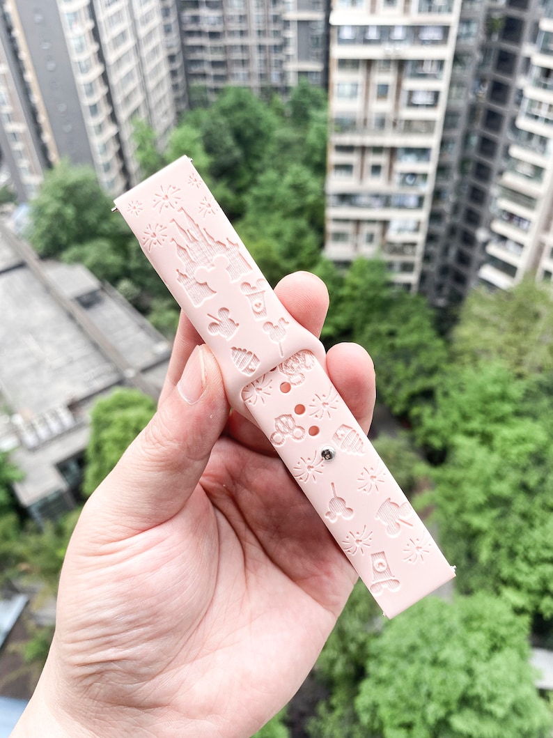 May include: A pink silicone watch band with a white embossed design featuring Disney characters and landmarks. The band has a small silver buckle.