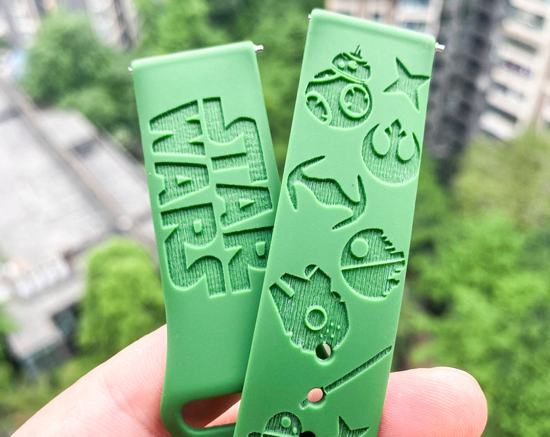 May include: Two green watch straps with Star Wars themed designs. One strap has the word "STAR WARS" embossed on it. The other strap has various Star Wars characters and symbols embossed on it, including R2-D2, a TIE fighter, and the Rebel Alliance symbol.