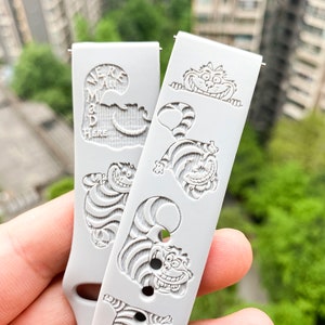 May include: Two light gray watch bands with engraved Cheshire Cat designs. One band has the text "We're all mad here". The bands feature various poses of the Cheshire Cat, with a smiling face and striped body.
