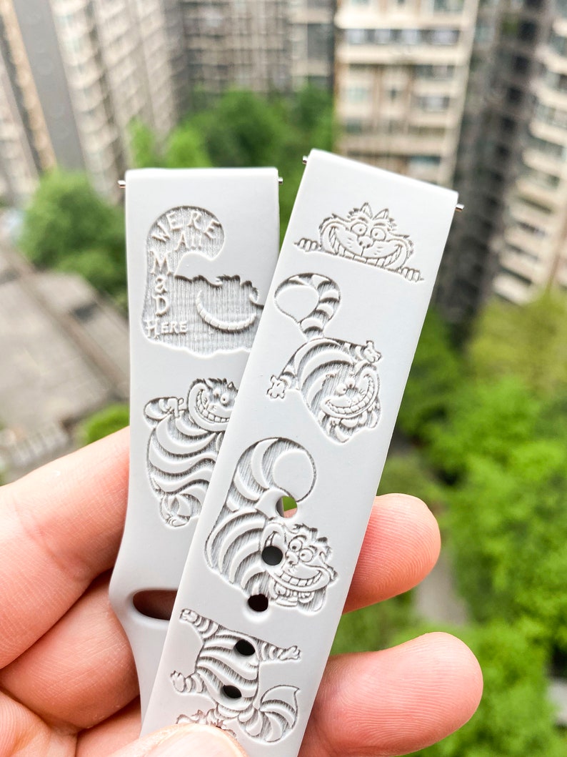 May include: Two light gray watch bands with engraved Cheshire Cat designs. One band has the text "We're all mad here". The bands feature various poses of the Cheshire Cat, with a smiling face and striped body.