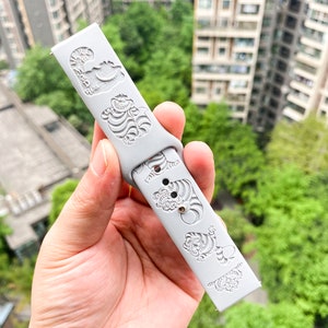May include: A light gray watch band with engraved designs of a smiling cat and tigers. The band is made of a flexible material and features a series of detailed illustrations. The watch band is held up against a background of green trees and buildings.