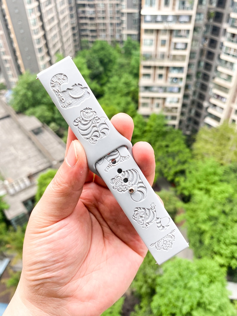 May include: A light gray watch band with engraved designs of a smiling cat and tigers. The band is made of a flexible material and features a series of detailed illustrations. The watch band is held up against a background of green trees and buildings.