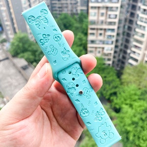 May include: A light blue silicone watch band with a repeating pattern of cartoon characters and designs. The band has a silver metal connector on one end.