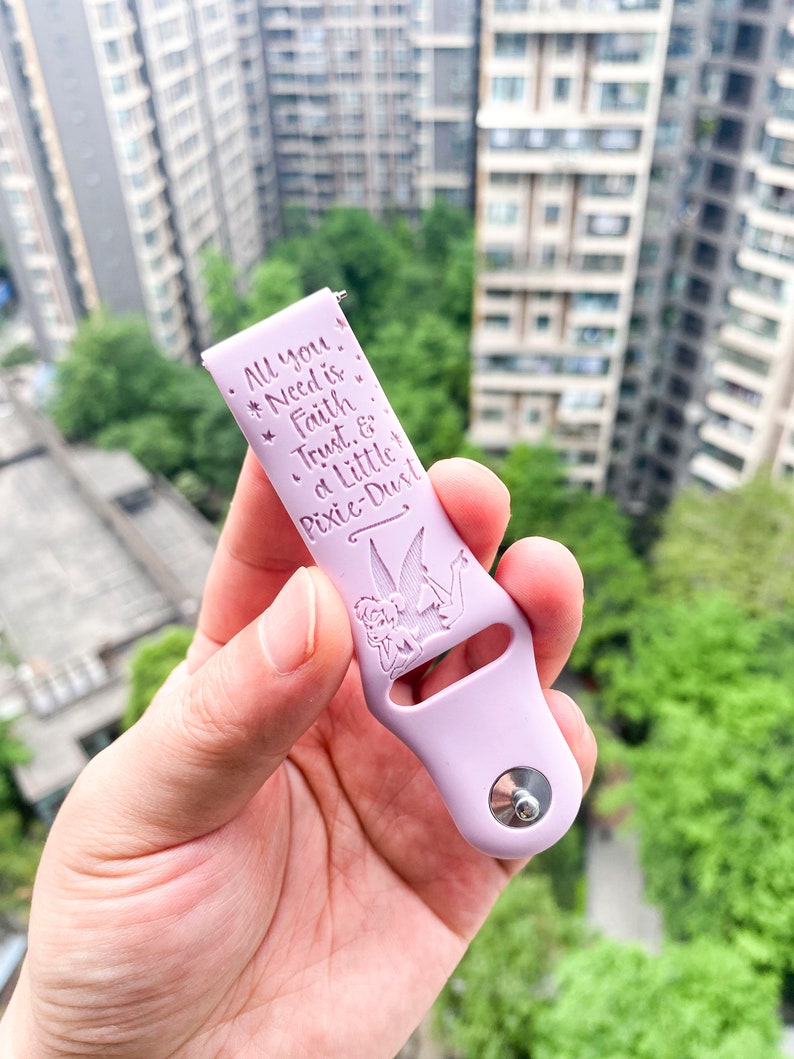 May include: A light purple silicone watch band with a silver metal clasp. The band has a Tinkerbell design and the text "All you *Need is *Faith *Trust & a Little Pixie-Dust".