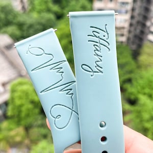 Könnte beinhalten: Zwei hellblaue Armbänder mit gravierten Designs. Ein Armband hat ein Herz und eine Herzschlaggrafik, das andere Armband hat den Namen "Tiffany" eingraviert.